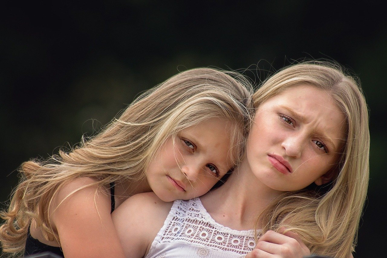Two girls with light hair share a close, affectionate hug outdoors; their faces are intentionally obscured.